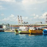 Embarcações atracadas em um porto industrial, com contêineres e guindastes ao fundo.
