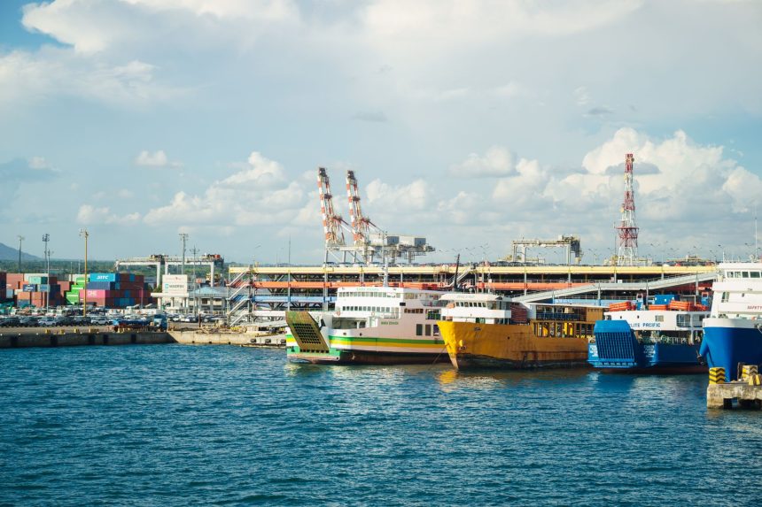 Embarcações atracadas em um porto industrial, com contêineres e guindastes ao fundo.