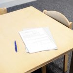 Documentos e uma caneta organizados sobre uma mesa próxima a cadeiras em uma sala clara.