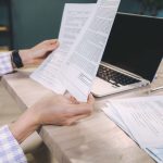 Mãos examinam documentos impressos ao lado de um notebook sobre uma mesa de escritório.